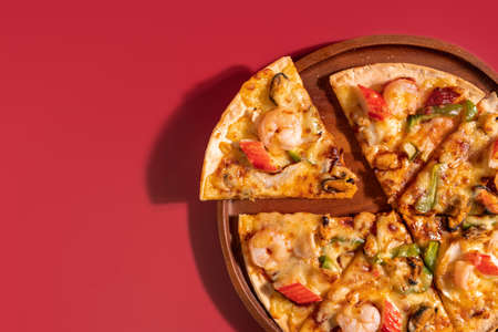 Close up slice of tasty hot baked seafood crispy pizza - mussels, shrimps and kani with chili pepper and melted mozzarella cheese on round pizza dough, on a wooden pizza pan over the red background.の写真素材