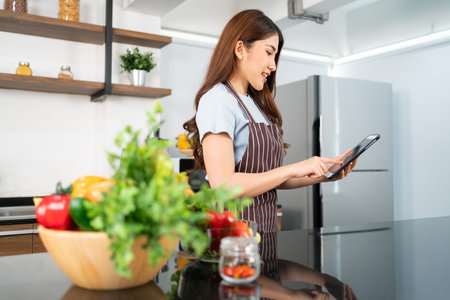 Happy Asian woman wearing an apron prepares vegetables for healthy food by searching recipes online on a digital tablet in home kitchen, watching videos of ingredients. Modern cooking technology.の写真素材