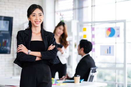 Portrait of a smiling professional Asian businesswoman wearing a black blazer standing in front of blur colleagues meeting in the background. Entrepreneur leadership image with copy space.の写真素材