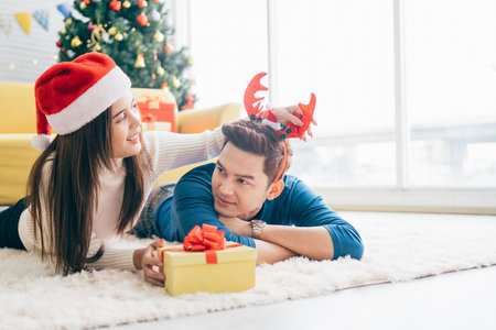 Young happy Asian woman wearing Santa Claus hat surprises her boyfriend with a Christmas gift at home while lay down on the carpet with Christmas tree in the background. Image with copy space.の写真素材