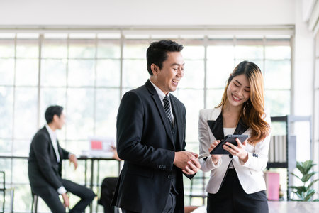 Senior Asian businessman and young female businesswoman discuss in the office, working on social media strategy, looking at a tablet with blur colleagues in the background. Image with copy space.の写真素材