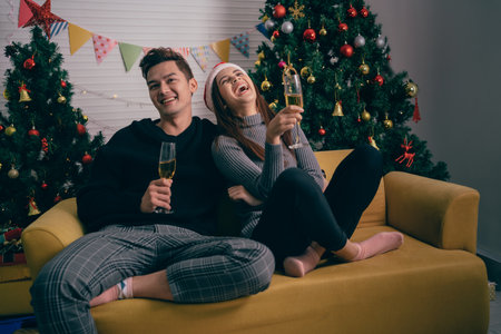 Happy Asian couple toasting champagne together, sitting on the sofa in the evening with a Christmas tree and lights in the background. Boyfriend and girlfriend at Christmas eve nightの写真素材