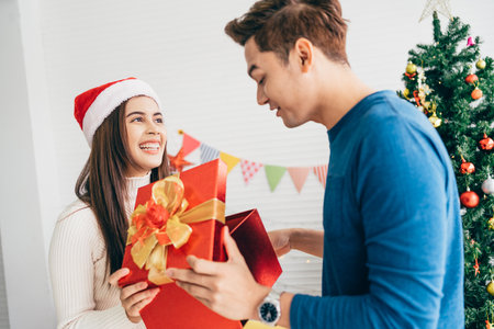 Young beautiful happy Asian woman wearing a Santa Claus hat surprises her boyfriend with gift at home with a Christmas tree in the background. Celebrate the boxing day concept. Photo with copy space.の写真素材