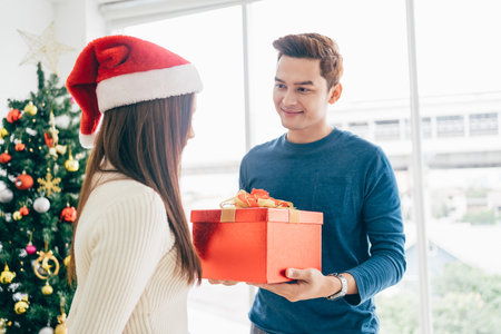 A happy Asian man surprises his girlfriend with Christmas presents at home with a Christmas tree in the background. image with copy spaceの写真素材