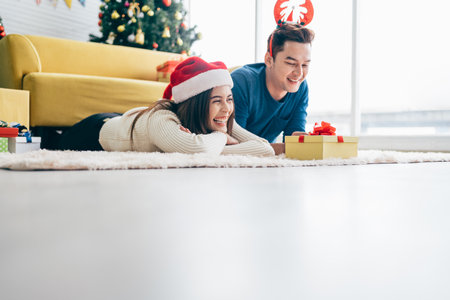 Young happy Asian woman wearing a Santa Claus hat with her boyfriend with a Christmas gift while lying down on the carpet with a Christmas tree in the background. Photo with copy space.の写真素材
