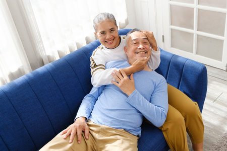 Portrait of happy Asian senior couple living together, hug, touching and embracing with a smile on the sofa in the living room. Retirement living together at home.の写真素材