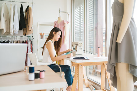 Portrait of a confident Asian tailor woman sewing clothes with a sewing machine. A young attractive female fashion designer smile and look at the camera with clothes rack in the background in studio.の写真素材