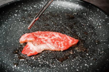 Premium Rare Slices sirloin Wagyu A5 beef with high-marbling texture pick up and grill on marble coated non-stick pan by bbq tongs with hand on black background. Served for Yakiniku.の写真素材