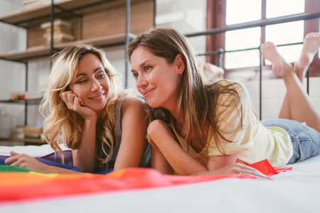 Happy Caucasian Lesbian couple looking at each other with a smile and love while laying together over the rainbow flag on the bed in the bedroom. LGBTQ couple spending quality time together concept.の写真素材