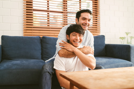 Happy caucasian and Asian LGBT couple, one sitting on the sofa embracing the other sitting on the floor from the back at home. Diversity of LGBT relationships. A gay couple concept.の写真素材