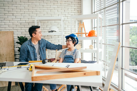 Cheerful Asian carpentry father discusses and teaches his son who wears a safety helmet about construction instruments. Construction work and repairs. Carpentry and family concept.の写真素材