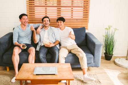 Gay people group sitting on the sofa, having popcorn and enjoying watching content on the tv in the living room. Three LGBT Caucasian and Asian people living together. Diversity of gay friendship.の写真素材