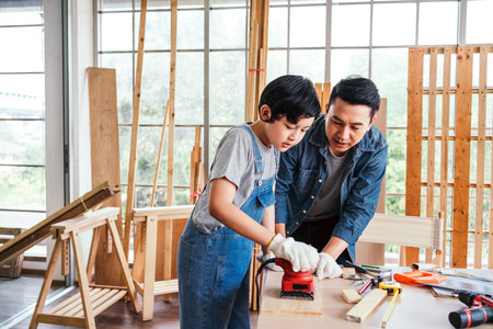Asian father and son carpentry sanding timber plank to smooth with an electric sander machine together carefully at the home carpentry studio. Carpentry working at a home workshop studio.の写真素材