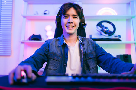 A young Asian man professional gamer wearing jeans jacket sits on a chair with a gaming table with pc, keyboard, monitor, microphone. Prepare for competition, cast gameplay or record a podcast.の写真素材