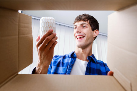 View from inside the box, a Smiling man unpacking awaited parcel, holding a vase, sitting with a...