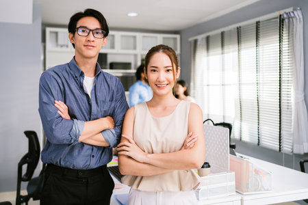 Cheerful young Asian businessman wearing eyeglasses and a businesswoman in casual smiling and looking at the camera while standing with her arms crossed. Happy businesswoman standing in the office.の写真素材