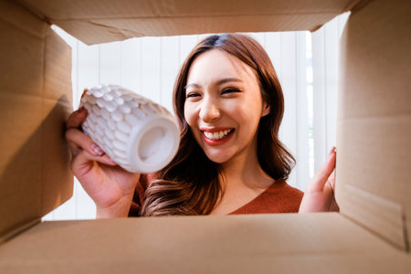 View from inside the box, a Smiling woman unpacking awaited parcel, holding a vase, sitting with a curtain in the background, satisfied happy customer opening a cardboard box with online store order.の写真素材