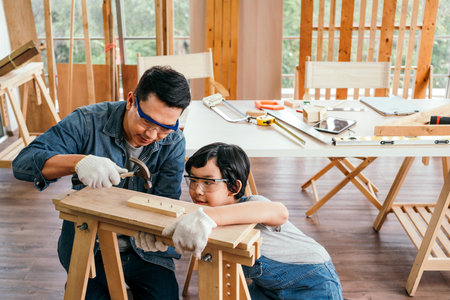 Happy Asian father and son work as a woodworker and carpenter. Father teaching his son to hammer nails on a wooden plank carefully together. carpentry working at a home workshop studio.の写真素材