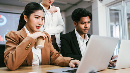 A confident Asian businesswoman holding a coffee cup, sitting and working on a laptop with her colleague with a woman standing in the back at her office. Asian businesswoman leader role at the meetingの写真素材