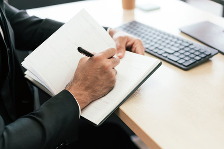 A closeup of the businessman's hands jotting something in a small notebook while looking at a computer monitor at his desk workplace. Businessman writing in the notebook while sitting in his workspaceの写真素材