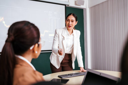 A cheerful and confident Asian businesswoman stands, present bar charts data from projector screen to her office colleagues. Asian business women leader role at the meeting.の写真素材