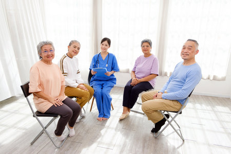 The caregiver therapist sits with a group of Asian senior people looks at the camera in a circle to check physical and mental health in a group elderly therapy session. The nursing home facilitatesの写真素材