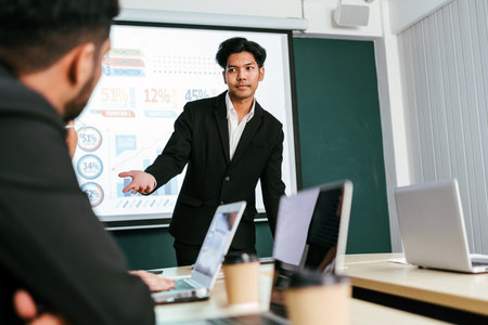 A cheerful and confident Asian businessman stands, presenting bar charts data from the projector screen to his office colleagues. Asian businessman leader role at the meeting.の写真素材