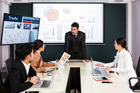 A cheerful and confident Asian businessman stands, presenting bar charts data from the projector...
