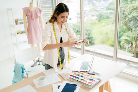 Asian woman fashion designer or tailor stands at the table, taking photos of fabrics and sample...