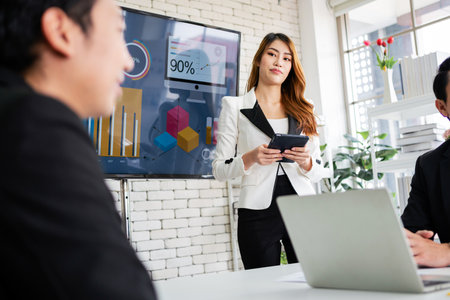 A cheerful and confident Asian businesswoman stands and uses a tablet to present a bar chart of data...