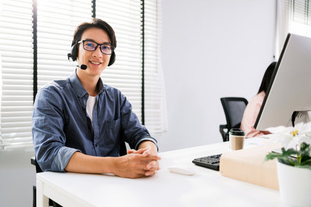A smiling Asian man in a headset sits at a desk in a bright office, ready to assist customers with professional support.の写真素材