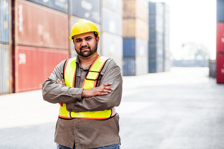 An Indian construction worker in a hard hat and safety vest poses confidently in a shipping yard...