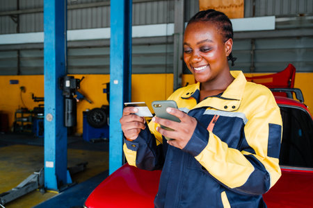 A smiling woman in a work uniform uses her phone and holds a credit card in a garage setting, showcasing a blend of technology and automotive work.の写真素材