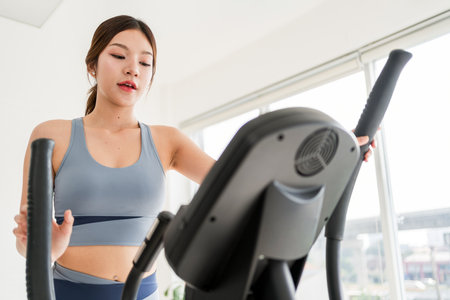 A focused young Asian woman exercises on an elliptical trainer in a bright, modern gym, promoting...