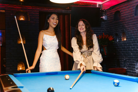 Two women enjoy a fun evening at a billiard table, laughing and playing pool in a stylish, dimly lit...