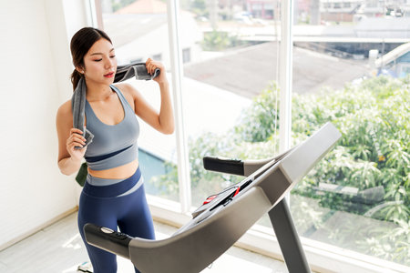An Asian young woman exercises on a treadmill in a bright room, holding a water bottle and looking...