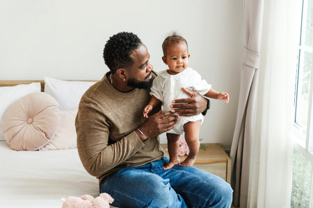 A loving father holds his baby daughter near a bright window, sharing a warm and tender moment. Cozy...