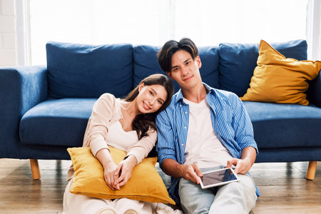 Young couple relaxes at home, leaning on a cozy blue couch. They smile while using a tablet, enjoying quality time. A warm, stylish living space reflects love, happiness, comfort, and modern lifestyleの写真素材