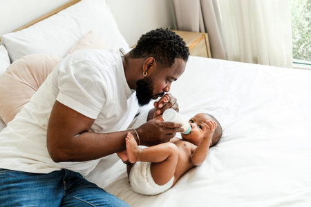 Loving African father bottle-feeding his baby in a cozy bedroom. Tender moment of bonding, care, and fatherhood. Parenthood, baby care, and family love concept in a warm, nurturing home setting.の写真素材