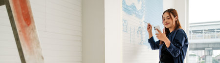 Young woman holding a phone and painting a wall with light blue designs during a DIY home decor project. Natural light streams through the window, enhancing the relaxed, creative vibe.の写真素材