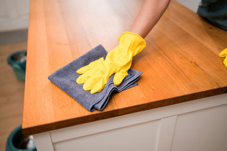 Close-up of a person in yellow gloves wiping a wooden kitchen counter with a dark cloth. Represents...