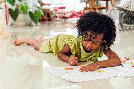 Cute Black girl in green dress lying on floor, coloring with crayons on large paper. Indoor creative...