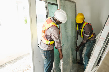 Two construction workers carefully install a large glass panel in an indoor site. Wearing safety helmets and vests, they collaborate on precise handling, showcasing modern building teamwork.の写真素材