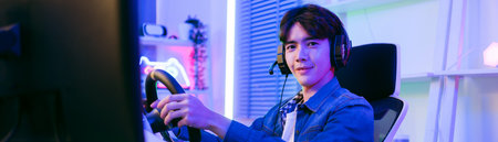 Smiling male gamer in headset and denim shirt plays a racing game using a steering wheel controller...