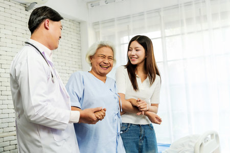 Smiling senior woman receives support from doctor and young caregiver, walking confidently in a hospital room. Depicts recovery, rehabilitation, healthcare, and emotional family connection.の写真素材