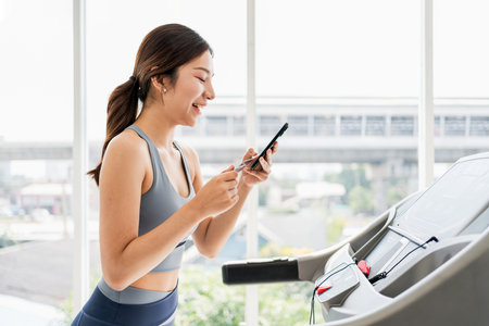 Young woman in active wear smiling while using smartphone on treadmill in modern gym. Healthy...
