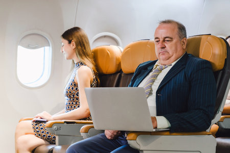 Businessman using onboard WiFi on a laptop while flying, seated beside a relaxed female passenger. Highlights inflight connectivity, digital services, and productivity in modern air travel.の写真素材