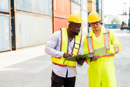 Two logistics workers wearing safety gear and hard hats coordinate container inventory using a tablet and clipboard at a busy port, representing teamwork in global shipping operations.の写真素材