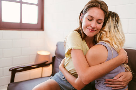Two women embrace warmly on a couch, eyes closed in comfort and love. A tender moment of LGBTQIA+ relationship support and affection in a peaceful, inclusive home environment.の写真素材
