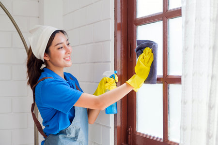 Smiling female housekeeper in apron and gloves cleaning a glass window with a spray and cloth. Professional home cleaning service promoting hygiene, neatness, and domestic maintenance.の写真素材